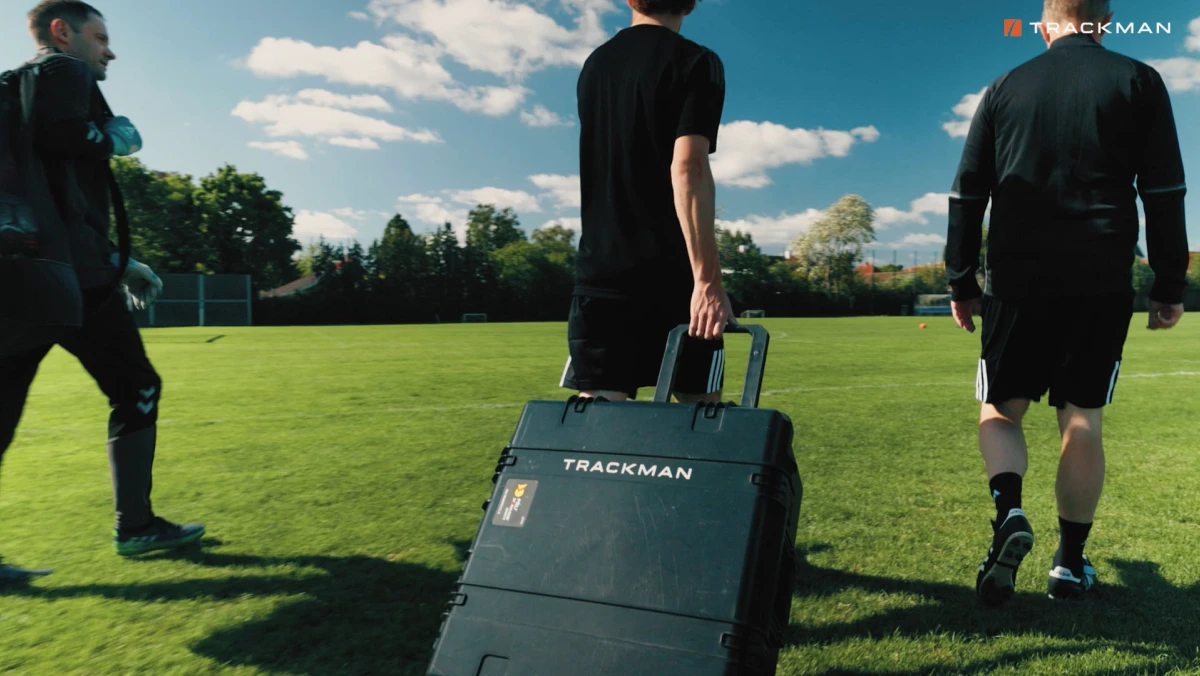Three people in sports attire walk on a grassy field, one pulling a "TRACKMAN" equipment case under a sunny sky.