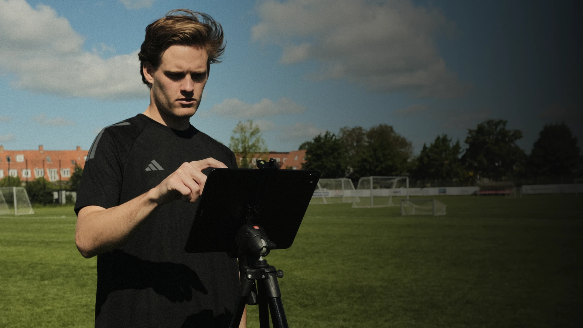 Man using a tablet on a tripod at a soccer field, with goals and trees in the background under a blue sky.