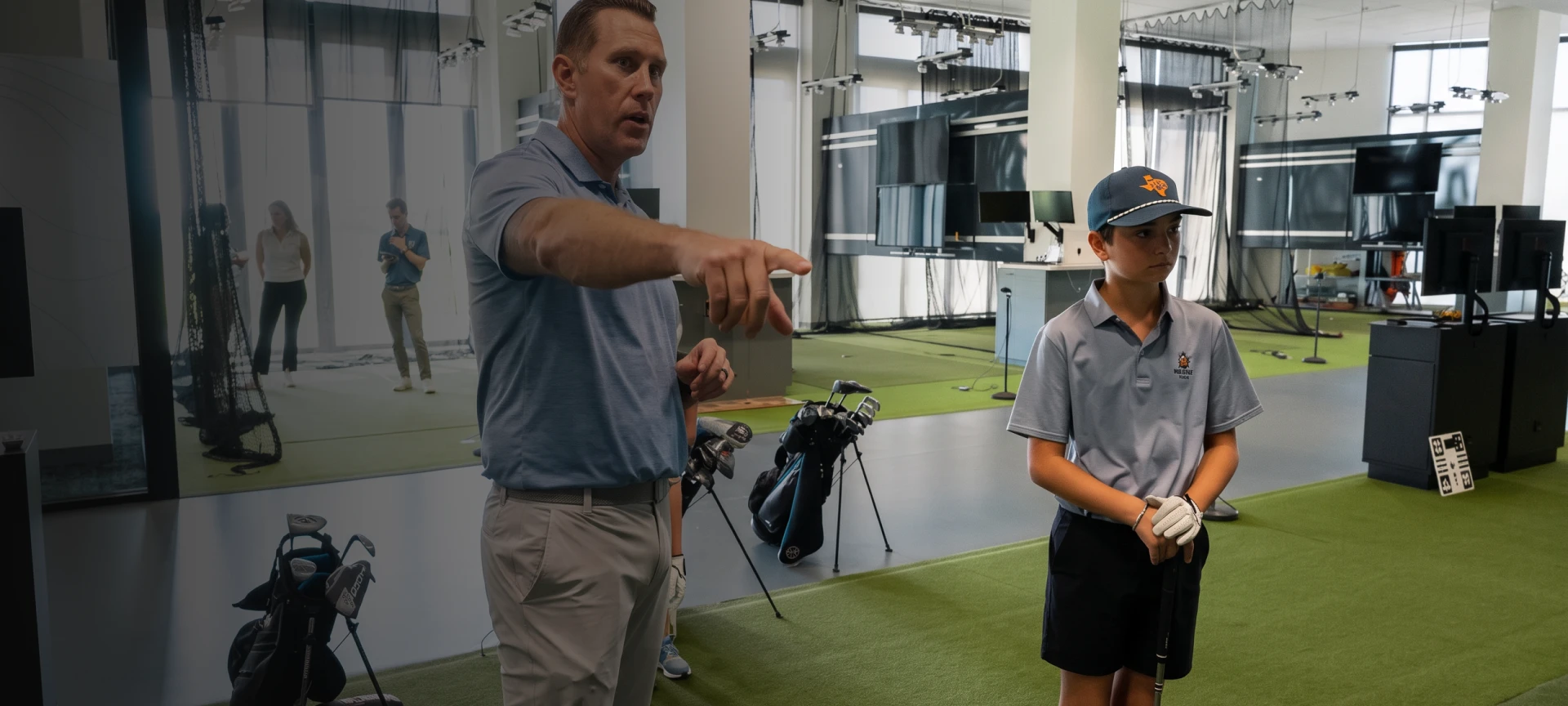 A man instructs a young boy holding a golf club inside an indoor golf training facility with golf bags and equipment in the background.