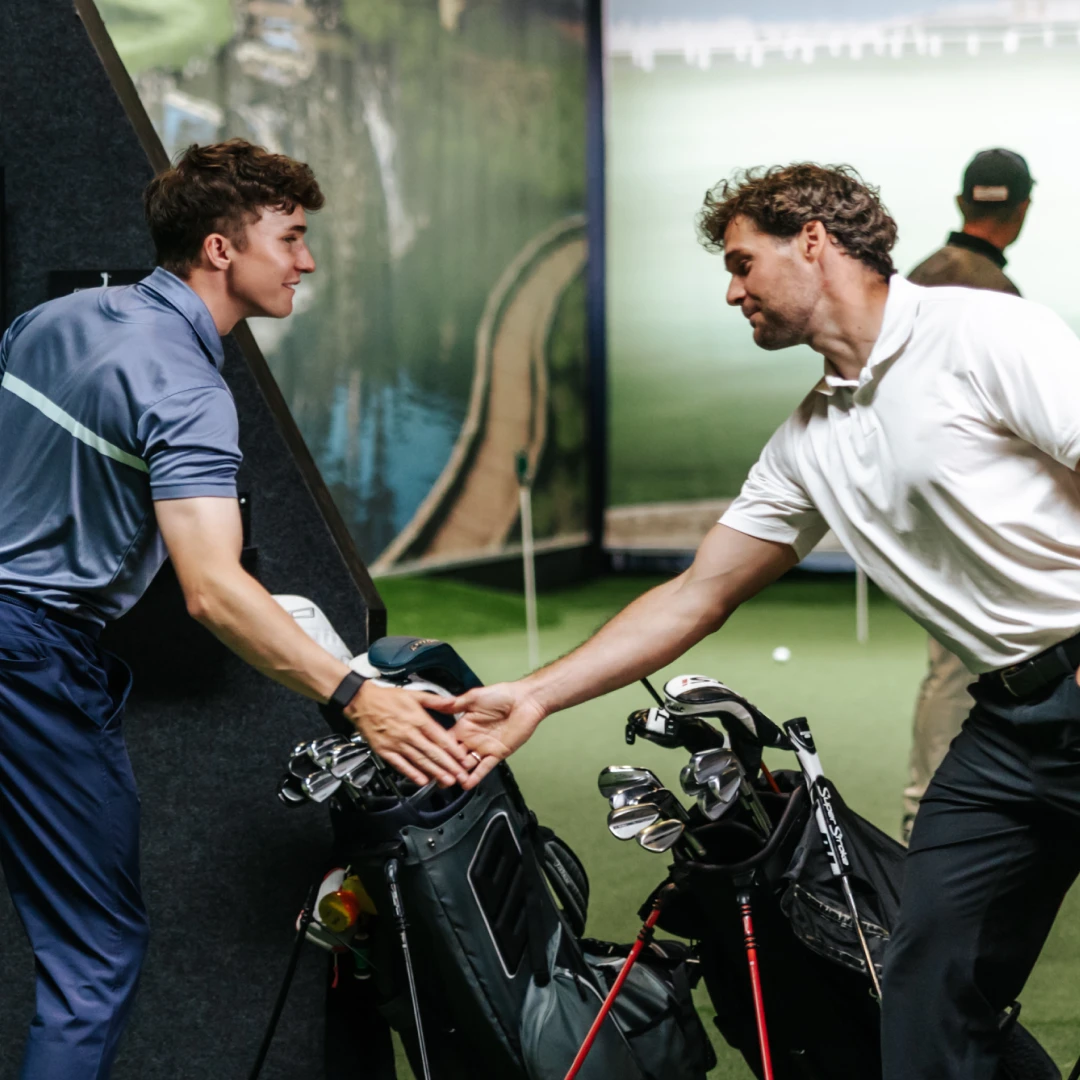 Two men shaking hands over golf bags indoors, dressed in casual golfing attire, with a golf course mural in the background.