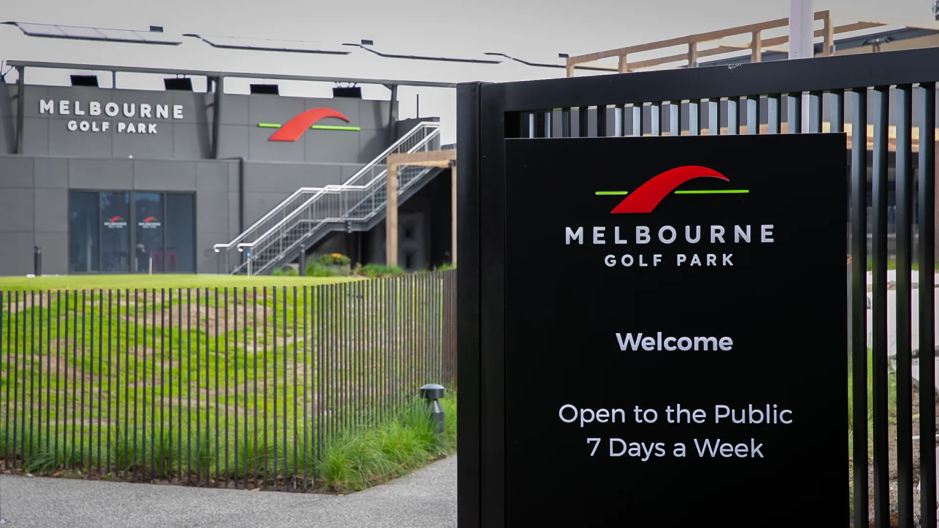 Black sign for Melbourne Golf Park, open to the public seven days a week, with a view of the main building and a grassy area.
