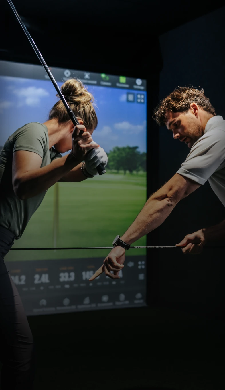 Two people practicing golf indoors with a simulator screen in the background, showing a virtual golf course.